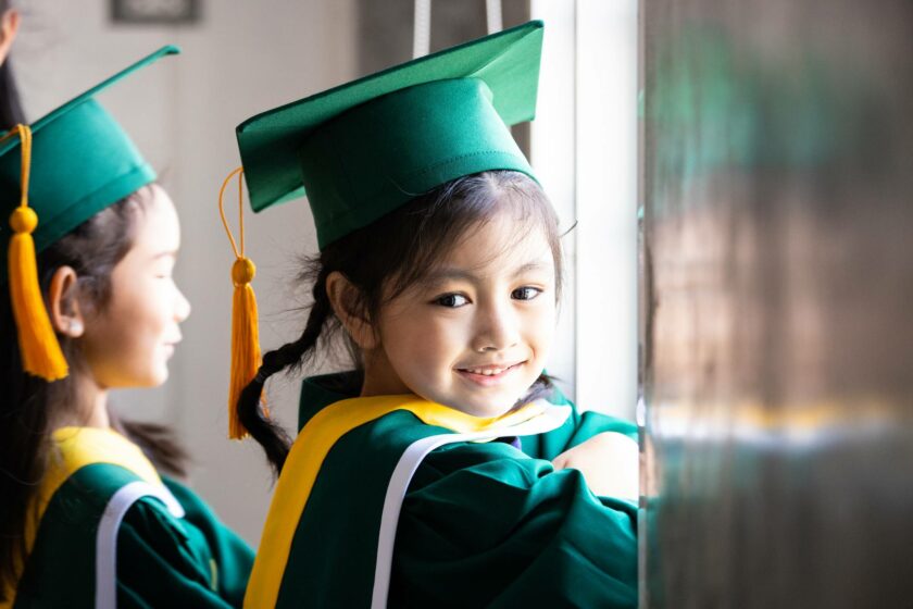 Two young girls in graduation gowns looking out of a window.